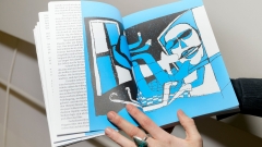 Eine Person hält ein aufgeschlagenes Buch in den Händen. Auf der rechten Doppelseite
                                          ist eine stilisierte Illustration in Blau, Schwarz und Weiß zu sehen: eine Figur sitzt vor einem Computerbildschirm. Die linken
                                          Seiten enthalten Fließtext. Die Hand der Person trägt einen auffälligen Ring mit türkisfarbenem Stein.