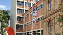 Eine Prideflagge vor der
                                          Universität für angewandte Kunst Wien. Darauf zu sehen ist ein Einhorn, ein Hippogreif und ein Phönix. Im Hintergrund gehen
                                          regenbogen-farbene Streifen ineinander über.