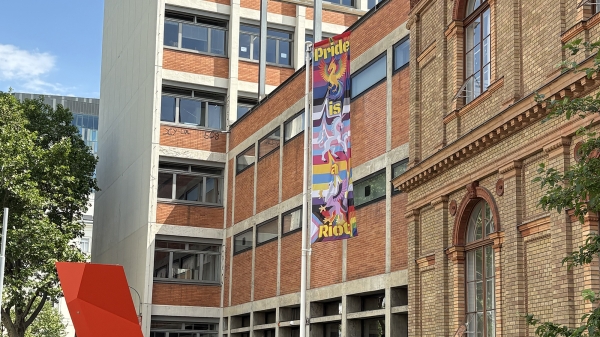 A pride flag in front of the University of Applied Arts Vienna.
                                          Background: Rainbow-coloured stripes. Foreground: A phoenix, a griffin and a phoenix.