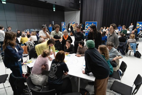 Eine Gruppe Menschen sitzt zu Gruppenarbeiten an Tischen zusammen, einige stehen bei Pinnwänden mit Post its
