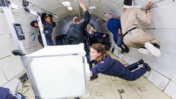 People in blue and beige flight suits float inside an aircraft cabin during a zero-gravity parabolic
                                          flight. Some hold onto the walls or equipment while others drift freely. In the foreground, one person interacts with a white,
                                          cube-shaped piece of equipment as the rest of the group moves in the background.