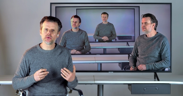 Ein Mann ist vier Mal in Bildschirmen zu sehen, und scheint einmal direkt mit dem Betrachter zu sprechen.