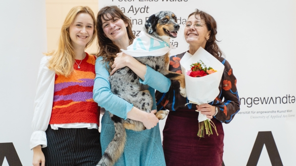Three women, one with a dog in her arms, in front of
                                          the lists of graduates.