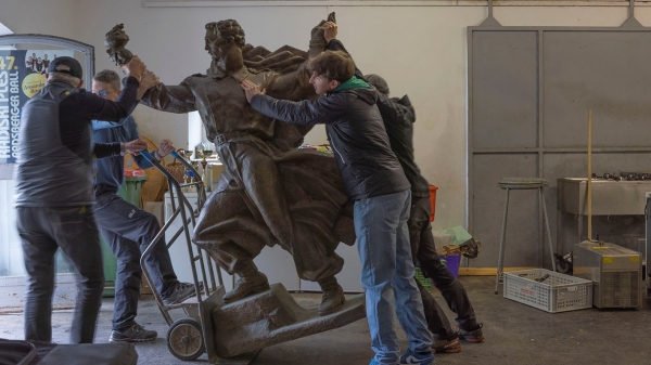 Ein Denkmal einer männlichen
                                          Figur wird von einer Gruppe Männer mit Sackkarre transportiert.