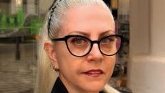 Fotoporträt ovn Basak Senova mit blondem, zurückgebundenem Haar und schwarzer Brille. Sie trägt ein schwarzes
                                          Sakko und eine Halskette und blickt direkt in die Kamera. Im Hintergrund ist ein Ausstellungsraum mit Glasflächen und Kunstwerken
                                          zu sehen.
