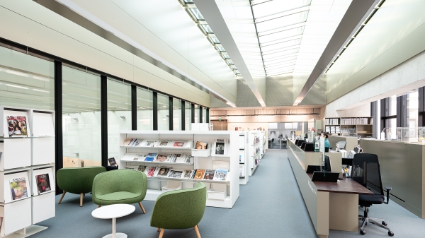 Interior view of the University Library at Vordere Zollamtsstraße
                                          7: bright reading area with modern shelves, green armchairs, magazine displays, and the information desk under a glass ceiling.
