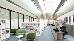 Innenansicht der Universitätsbibliothek
                                          in der Vorderen Zollamtsstraße 7: heller Lesebereich mit modernen Regalen, grünen Sesseln, Zeitschriftenauslage und Informationstheke
                                          unter einem Glasdach.