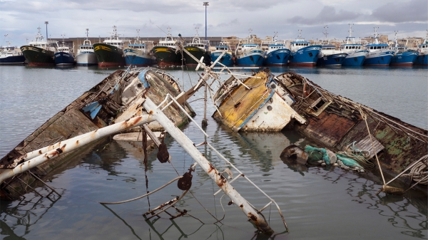 Plakat zur Ausstellung „Soil &
                                          Water: Mediterranean Crossing“ mit einem Foto von versunkenen, beschädigten Fischerbooten im Vordergrund und einer Reihe intakter
                                          Boote im Hafen im Hintergrund. Das Foto stammt aus der Serie Hotspot Mediterraneo (2025) von Francesco Bellina. Courtesy of
                                          the artist.