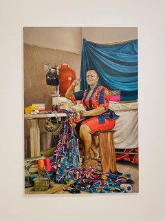 Das Ölbild von Afia Prempeh zeigt eine schwarze Schneiderin in einer Schneiderei. Sie schaut das Publikum direkt an.